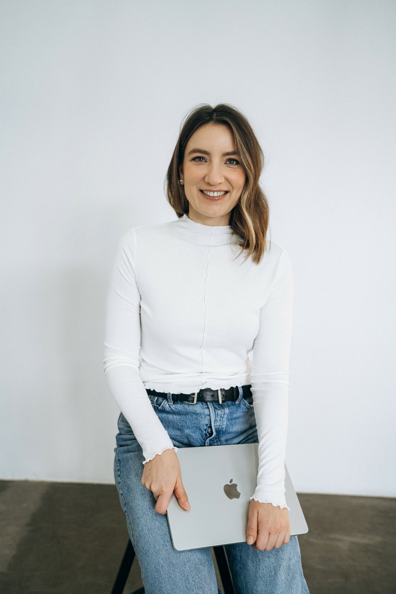 Smiling woman with shoulder-length hair holding a laptop, wearing a white turtleneck and jeans, standing against a plain background.