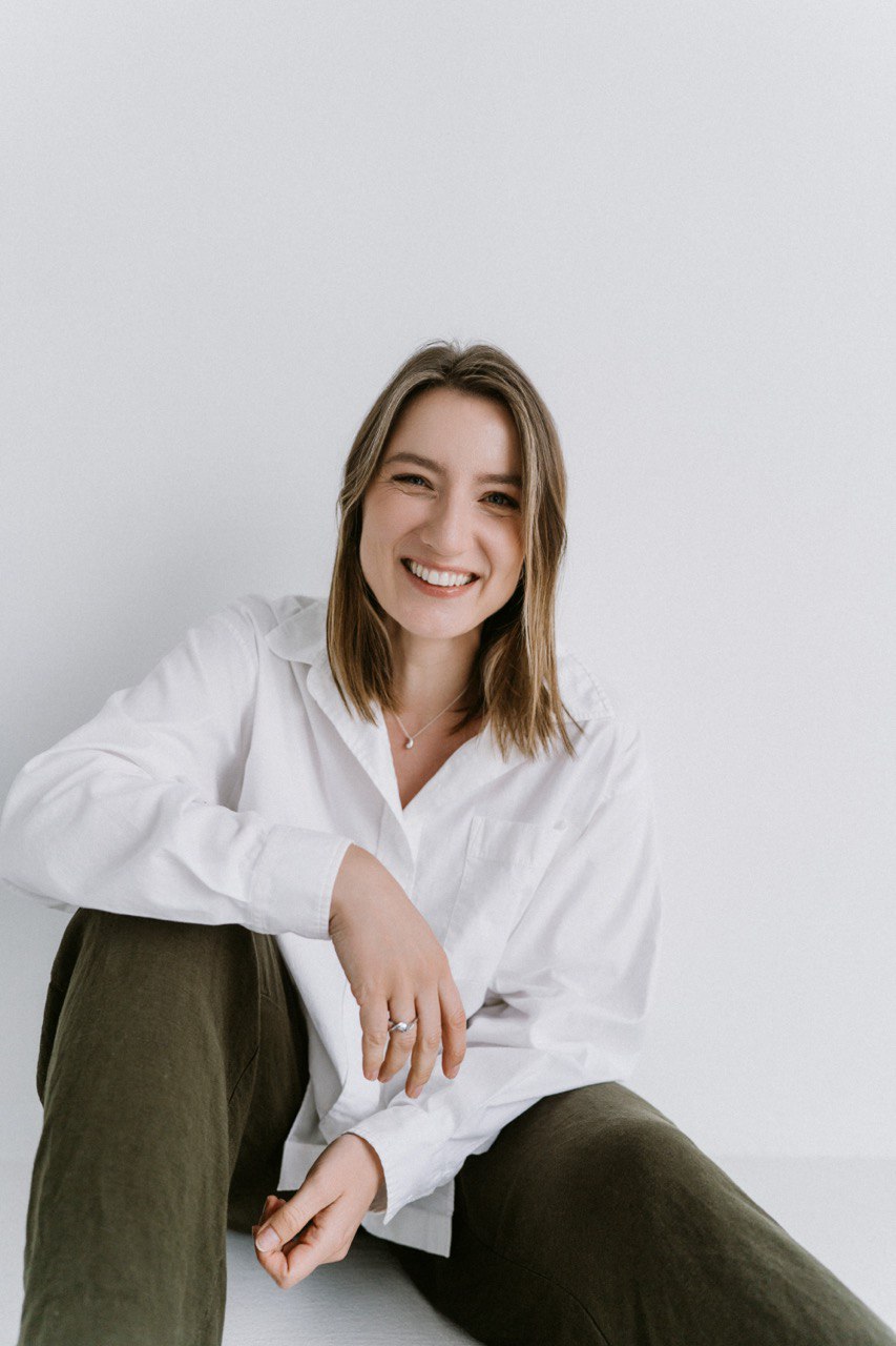Young woman with shoulder-length hair smiling, wearing a white shirt and dark pants, sitting against a plain white background.