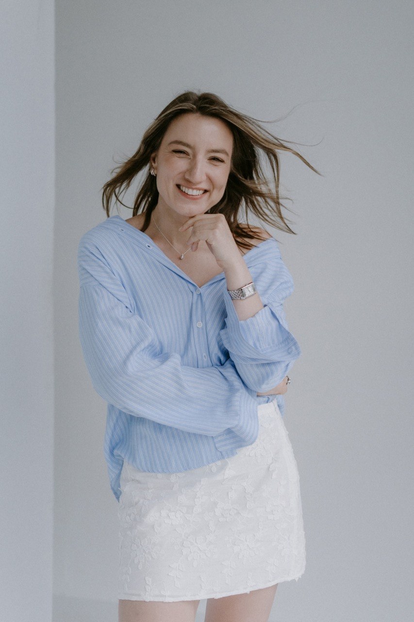 Smiling woman with shoulder-length hair, wearing a light blue striped shirt and white skirt, standing against a plain background.