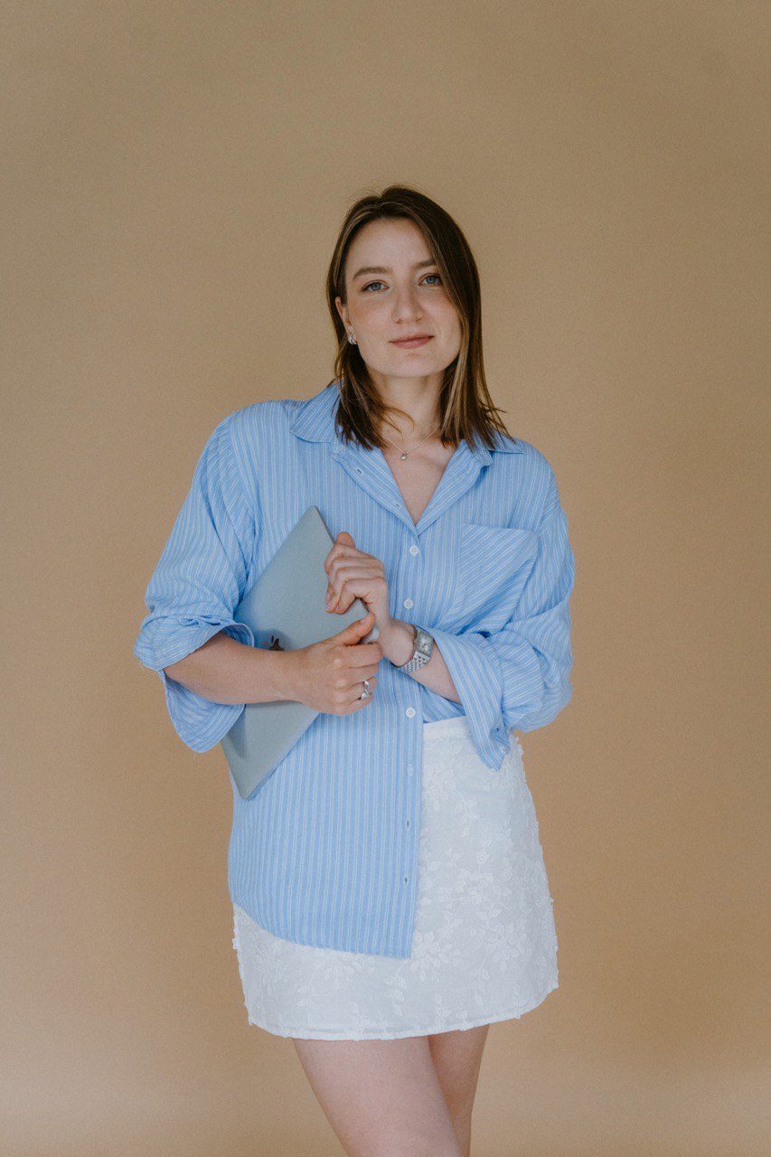 Young woman with shoulder-length hair wearing a blue striped shirt and white skirt, holding a phone, standing against a beige background.