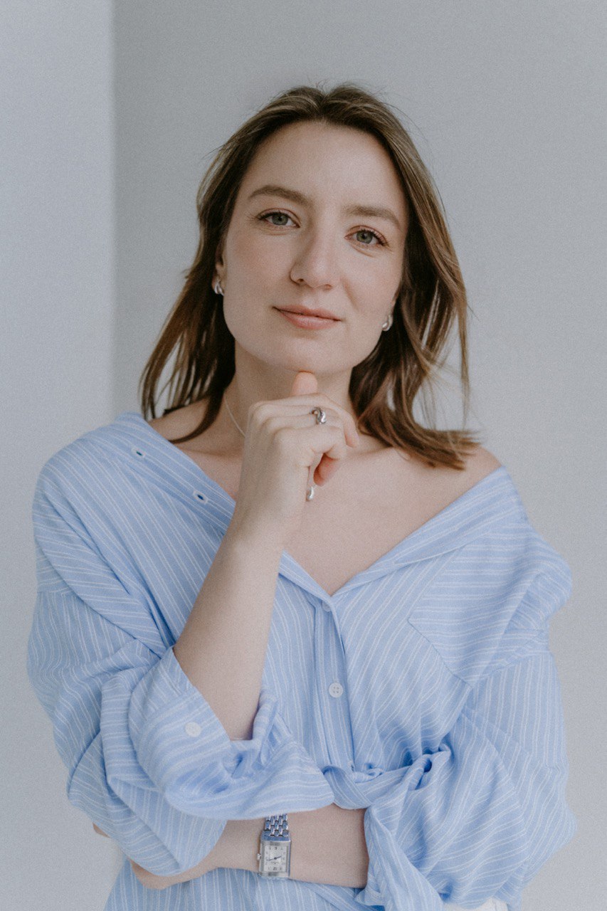 Young woman with shoulder-length hair, wearing a light blue striped shirt, standing against a plain background.