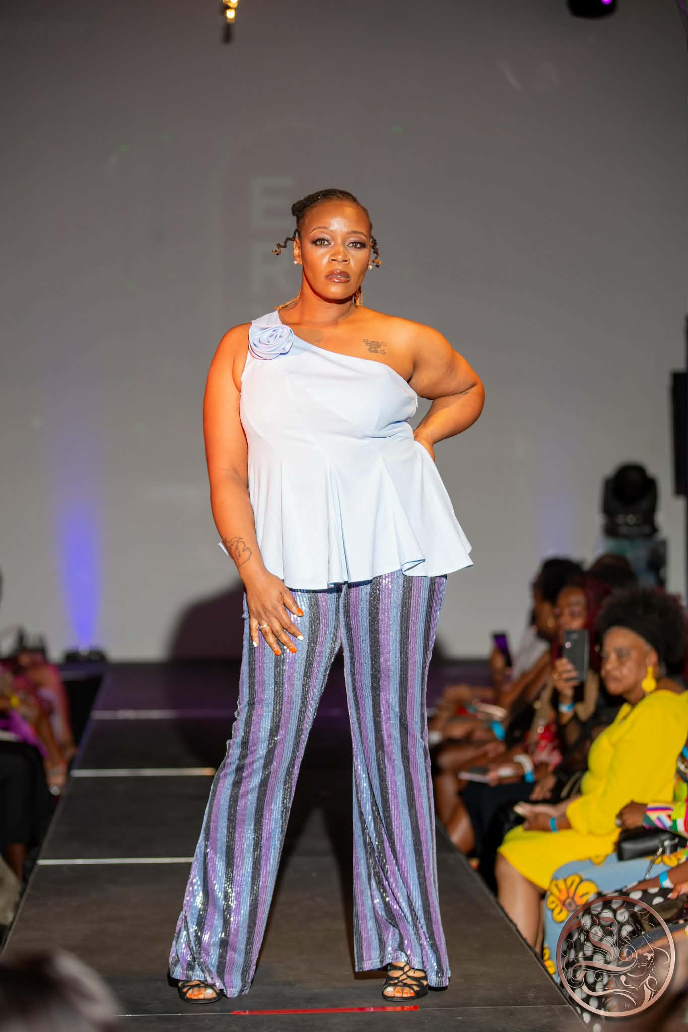Woman walking on runway in colorful striped pants and white top, audience seated on sides, plain background.