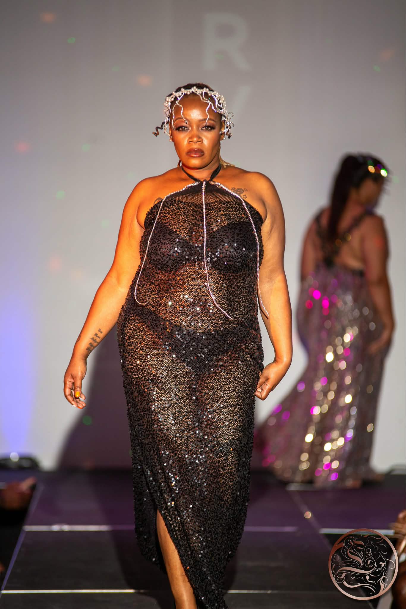 Woman walking on runway in black sequined dress with slit, headpiece, and jewelry, with another person in sequined outfit in background.