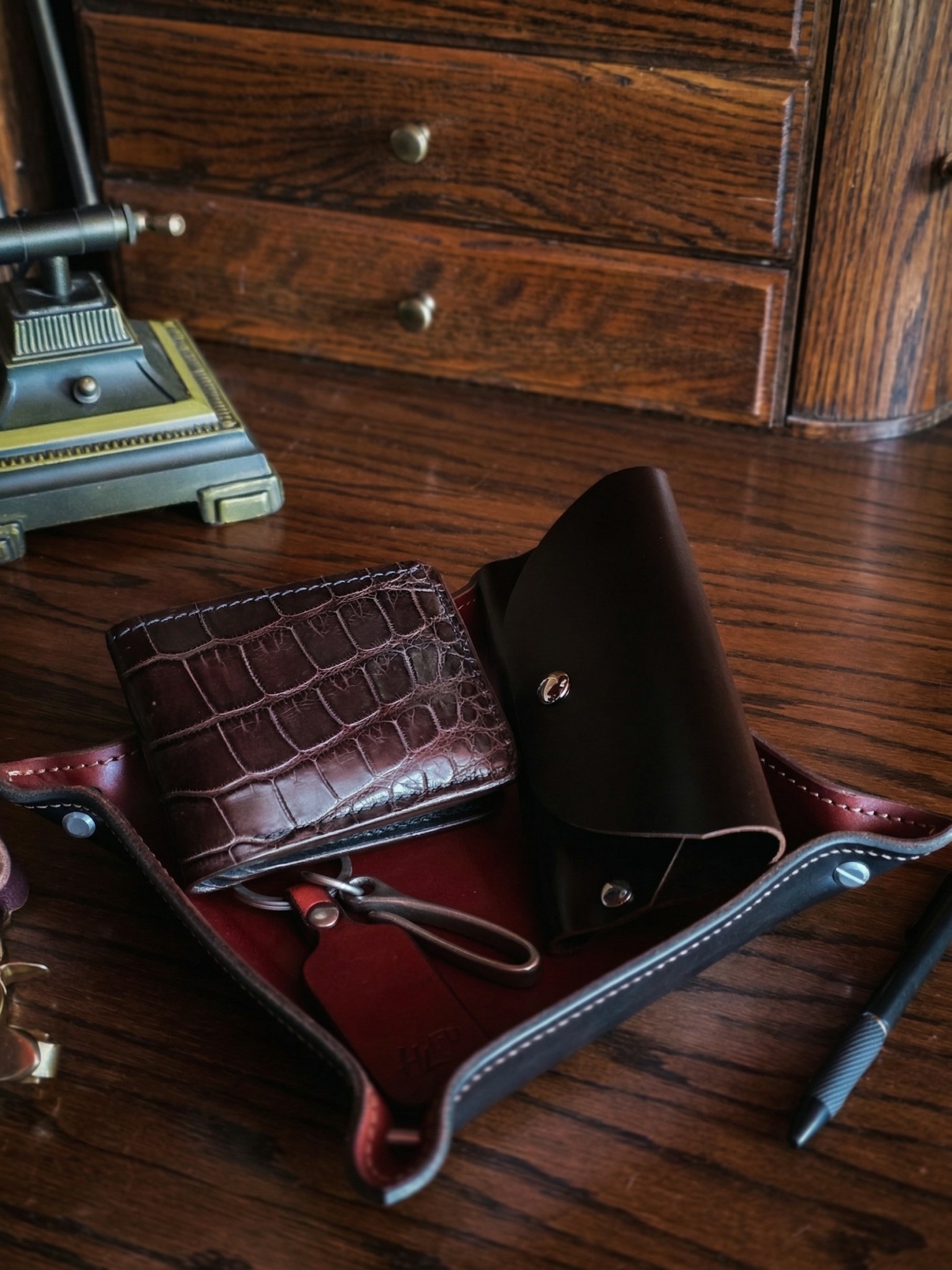 Open leather bag with wallet, flashlight, and pen inside, on wooden floor near wooden drawers.