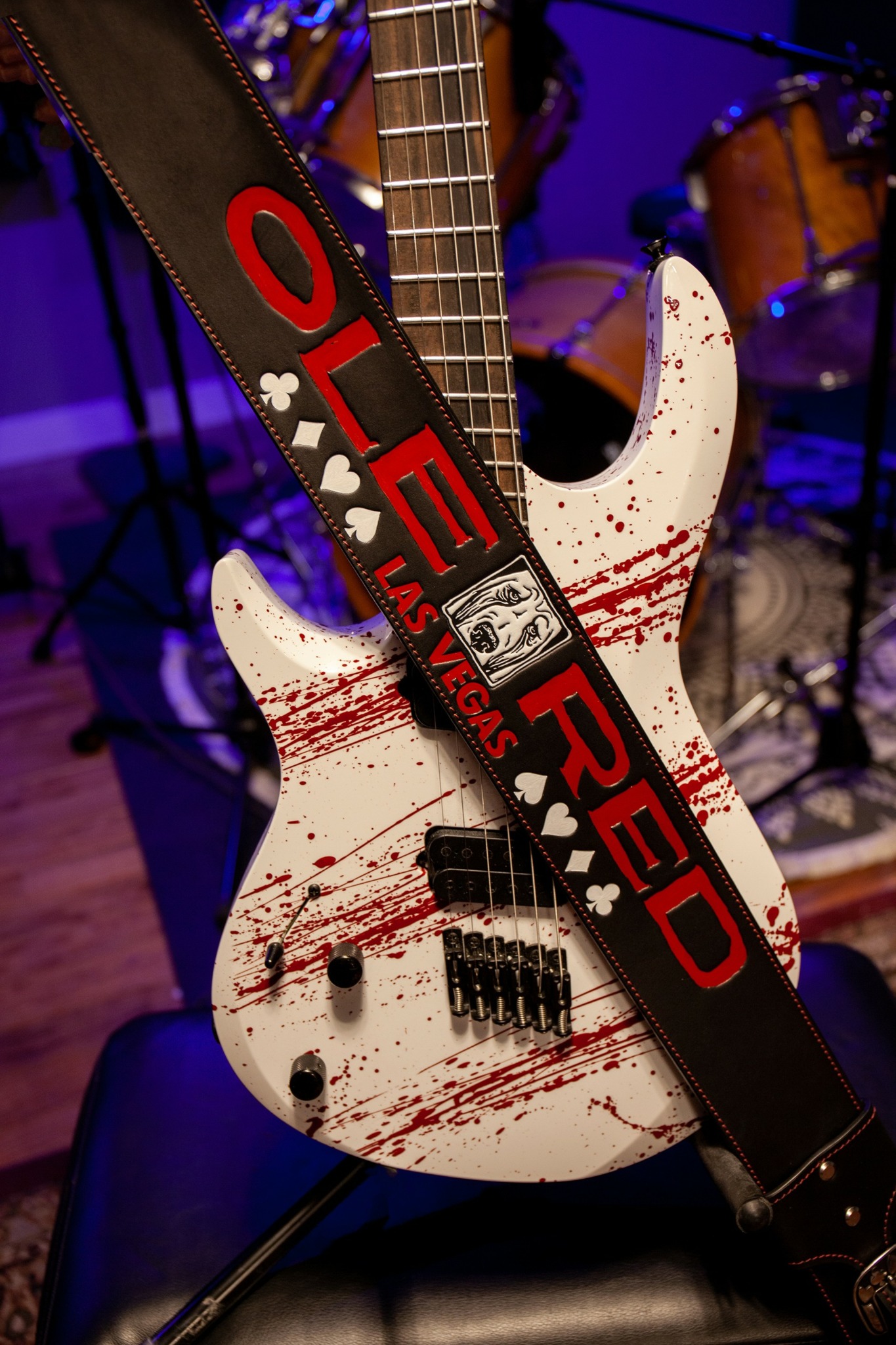 Electric guitar with blood-like red streaks, strap with 'LOVE HATE' text, background with drums, on a stool.