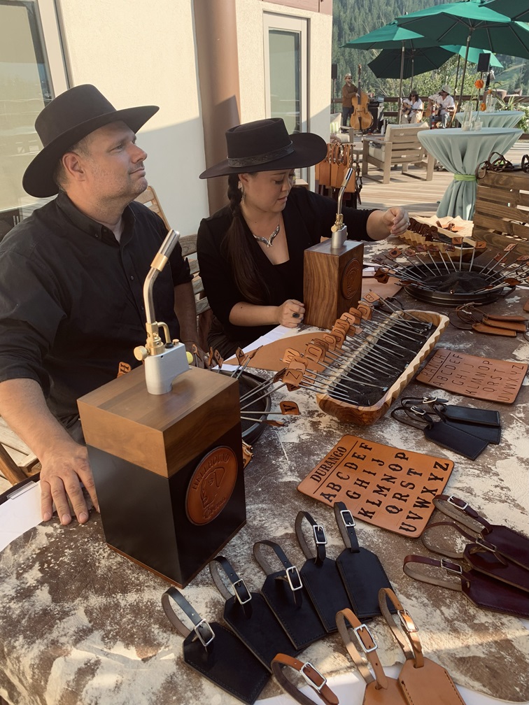 Two people sit at a table with vintage microphones, a wooden box, and various items, outdoors with umbrellas and people in background.