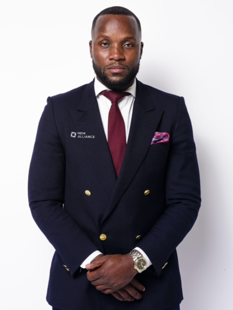 Man in a dark suit with a white shirt, maroon tie, and pink pocket square, standing against a white background.