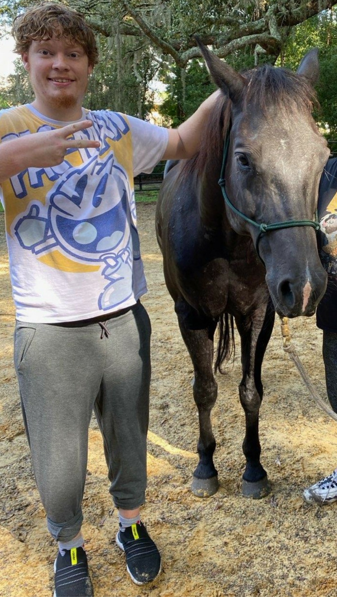 Person with curly hair in a white T-shirt and gray pants standing next to a horse outdoors.