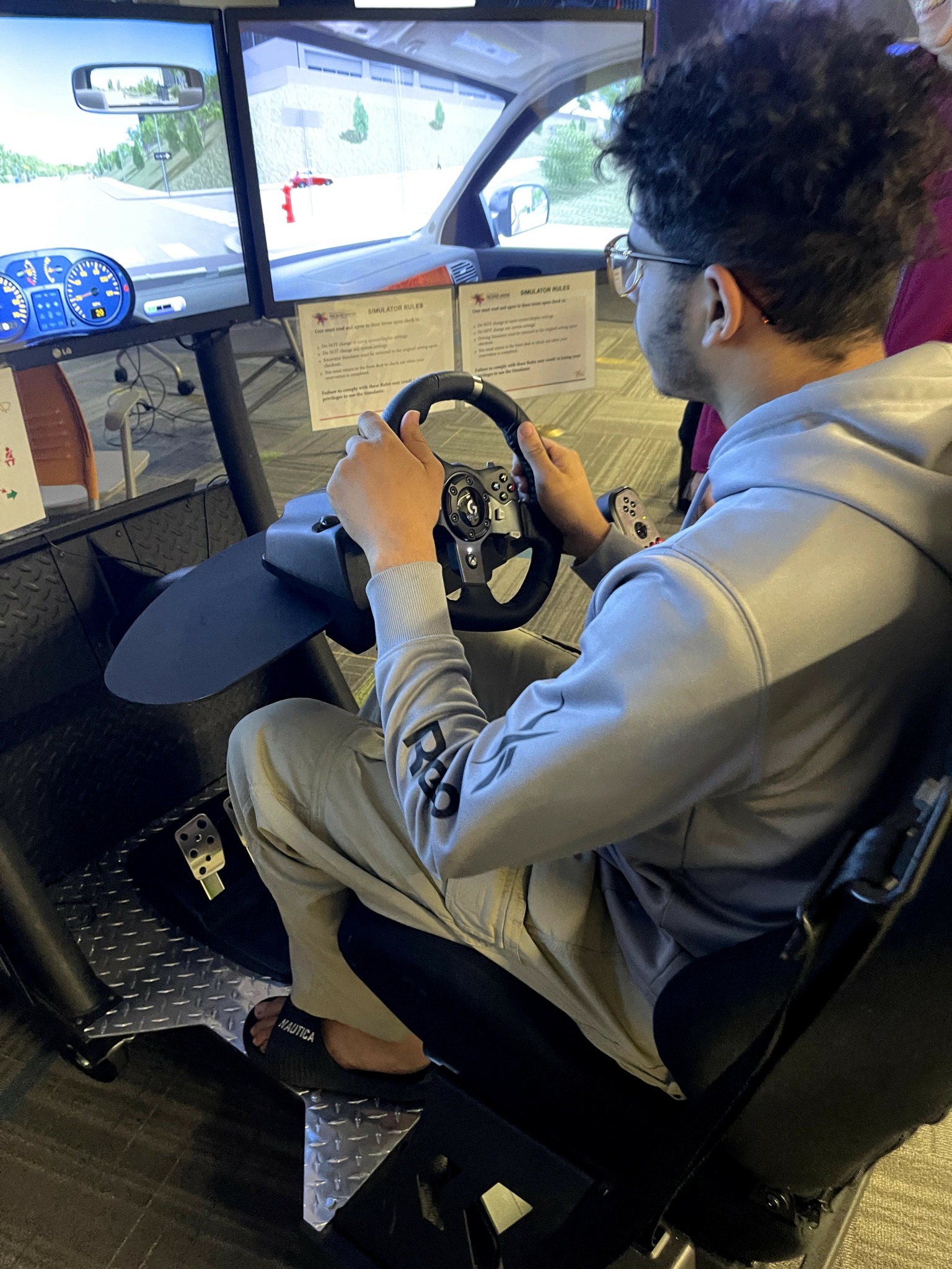 Person with curly hair and glasses sitting in a driving simulator, holding a steering wheel, with multiple screens displaying road views.