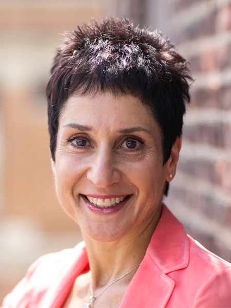 Smiling woman with short dark hair, wearing a pink blazer, standing outdoors near a brick wall.