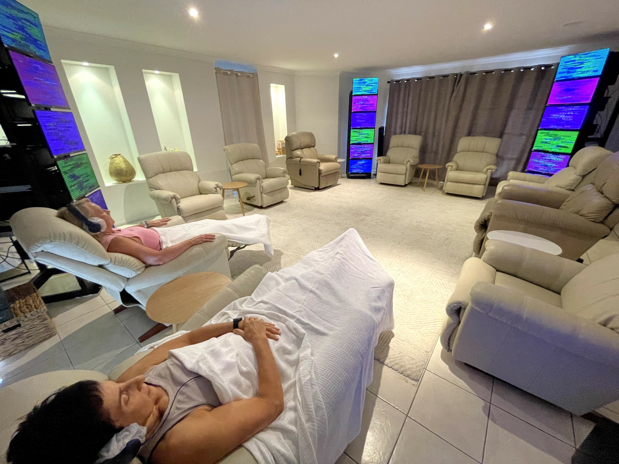 Person lying on a hospital bed in a room with multiple chairs and colorful vertical screens, bright lighting, and beige furniture.