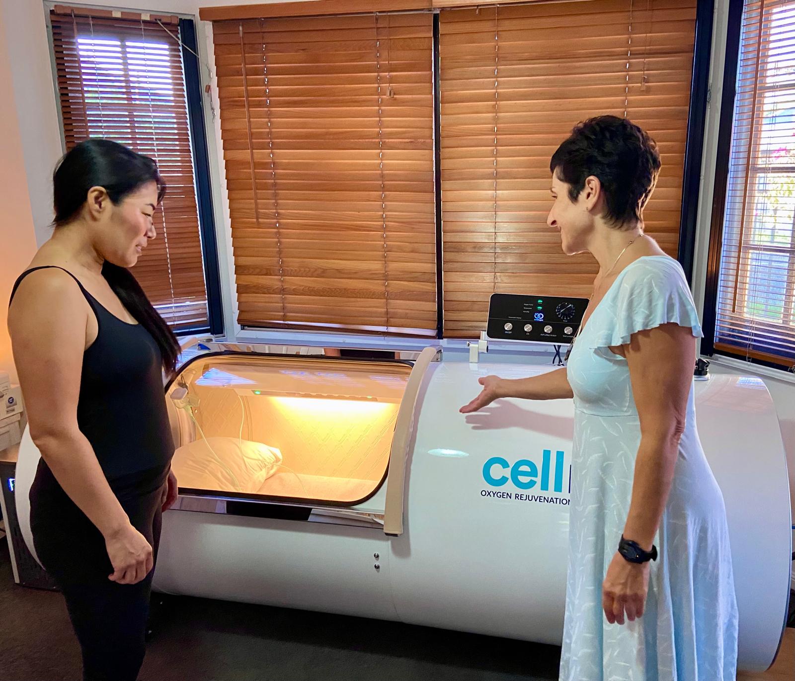Two women standing next to a large medical device in a room with wooden blinds.