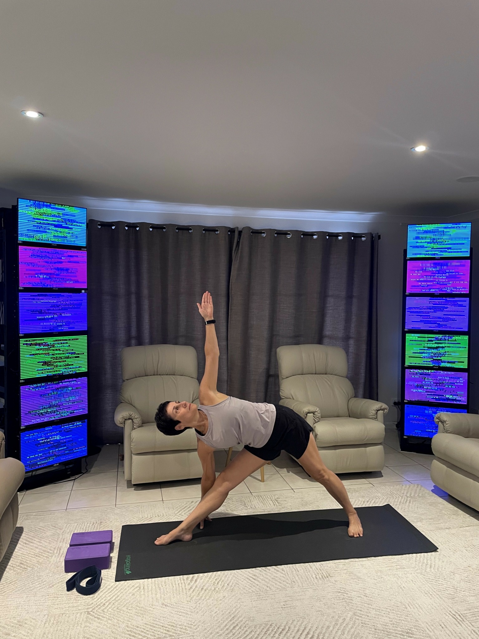 Person performing a yoga pose on a mat in a living room with two armchairs and two tall screens displaying colorful text.