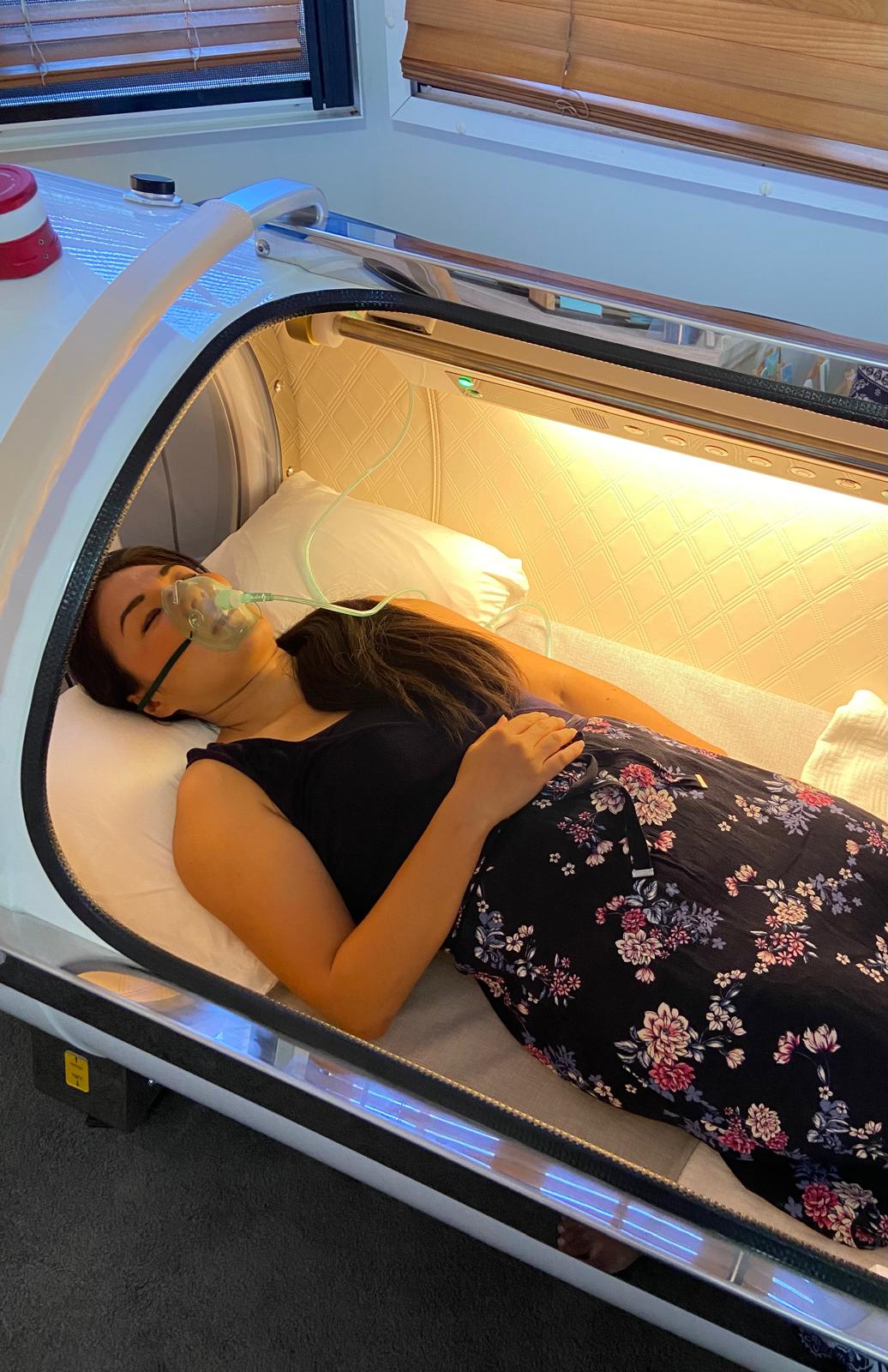 Person lying inside a medical imaging machine, with eyes closed and breathing mask, in a room with wooden blinds.