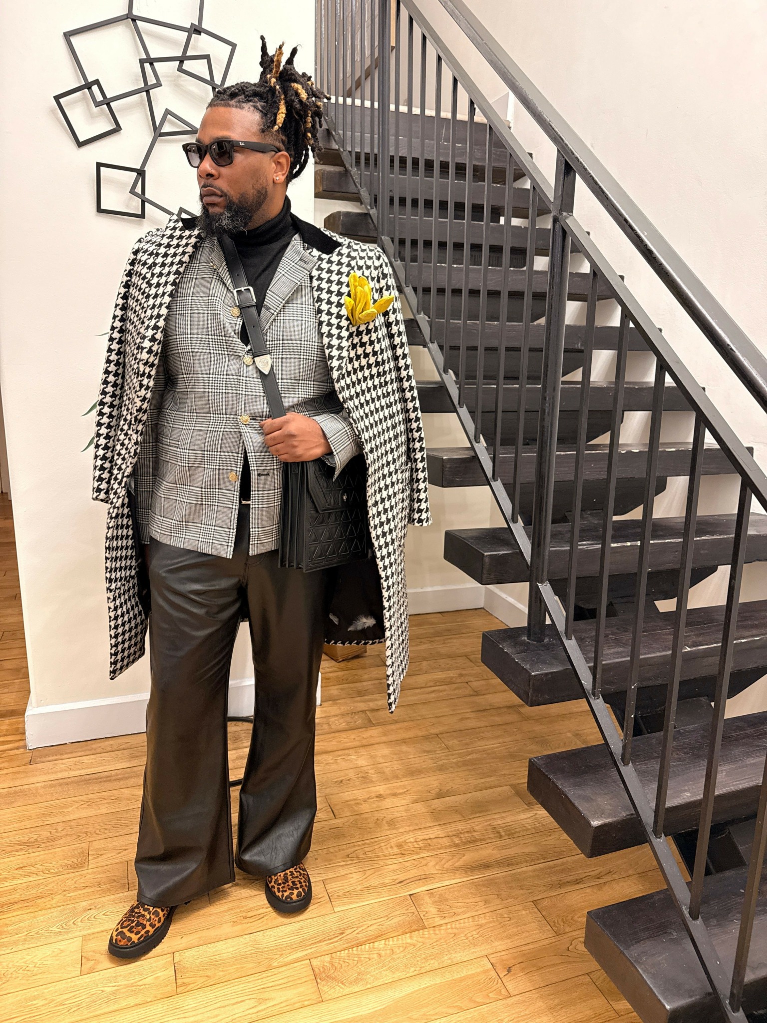 Man with sunglasses and dreadlocks stands near staircase, wearing a checkered coat, gray blazer, black pants, and patterned shoes.
