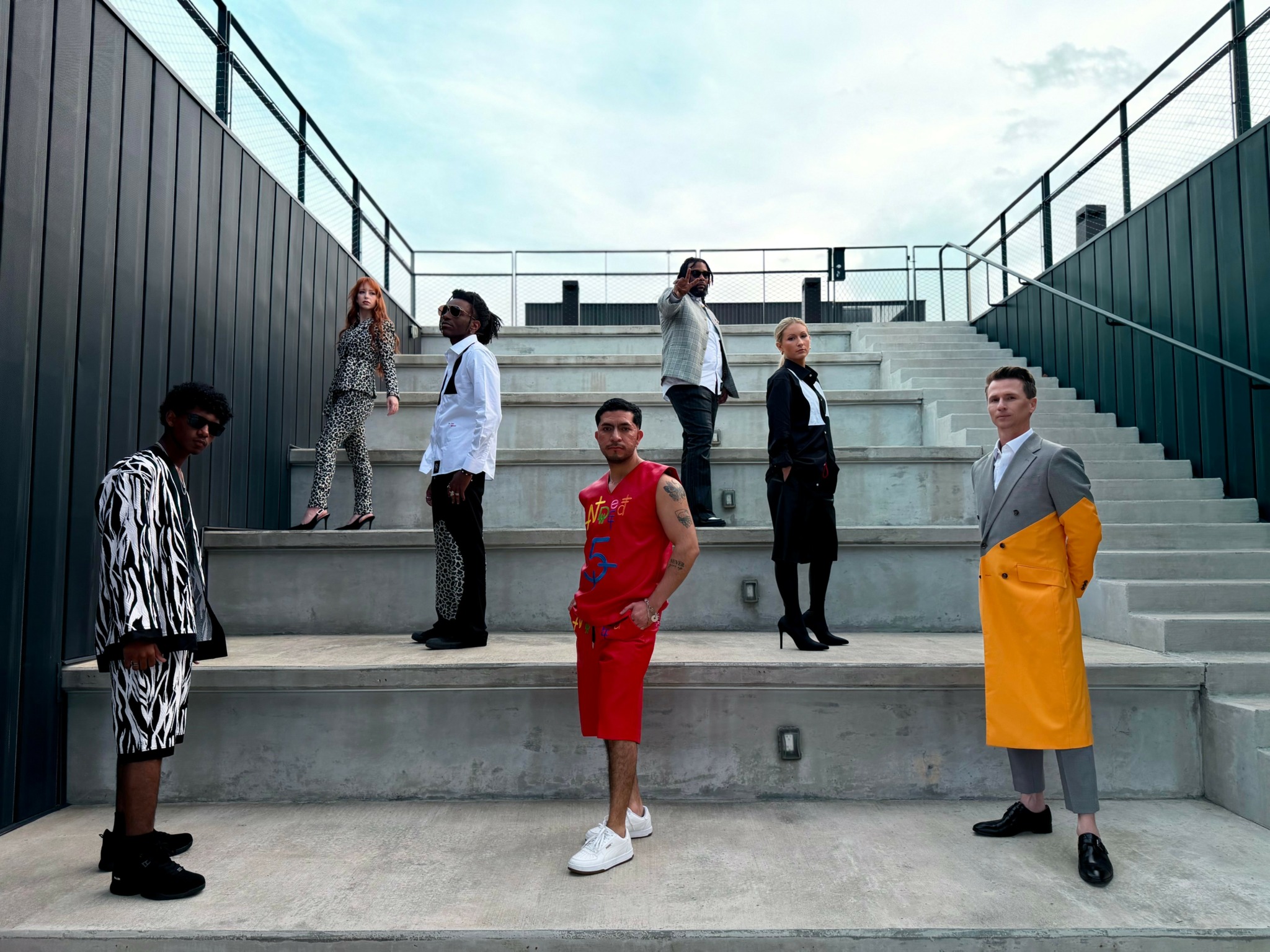 Seven people standing on concrete steps outdoors, some looking at the camera, others looking away, with a metal railing and sky background.