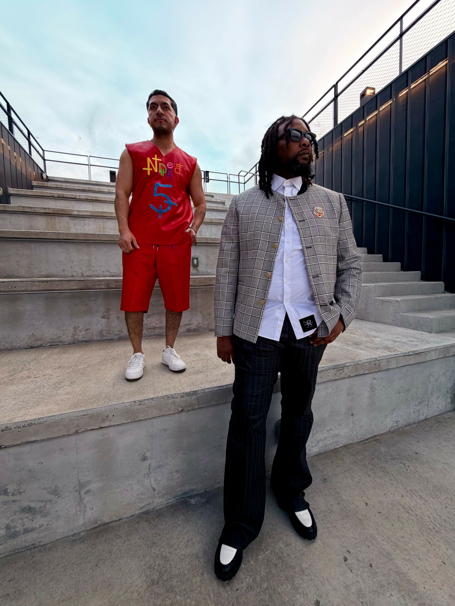 Two men standing on concrete stairs outdoors, one in a gray blazer and black pants, the other in a red sports outfit.