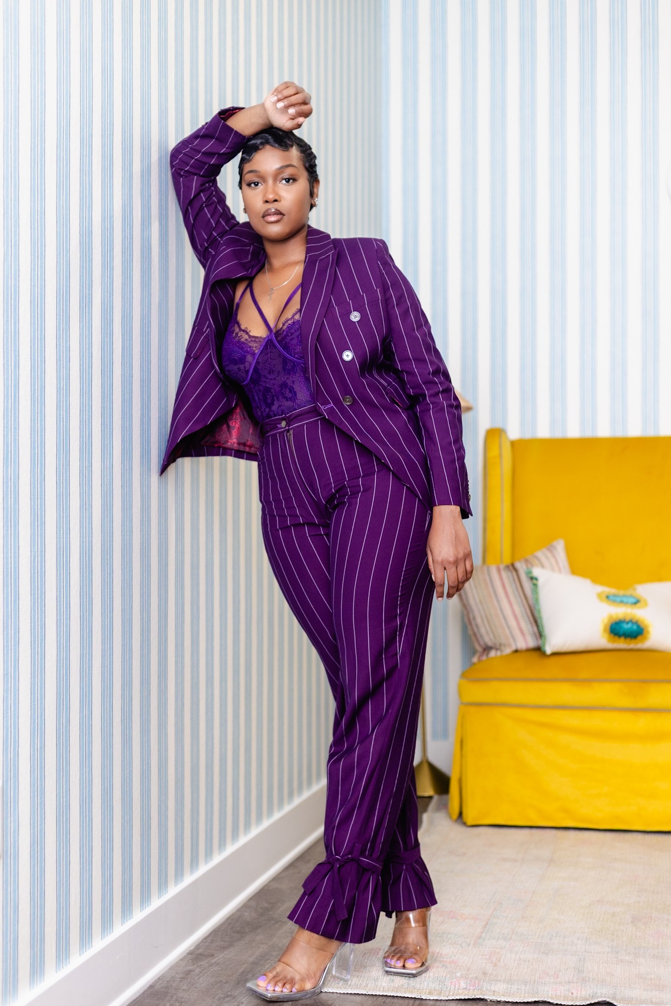 Woman in purple striped suit standing next to yellow sofa with pillows, one hand on head, in room with striped wall.