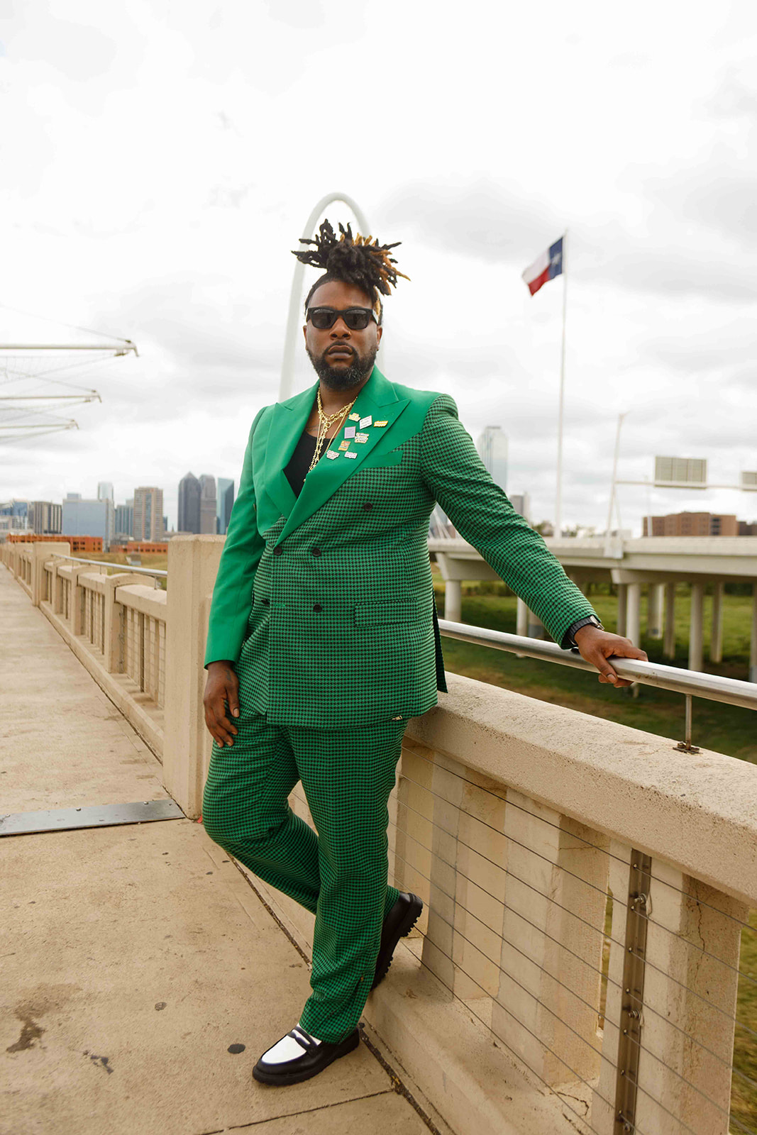 Man in green suit with sunglasses standing on a bridge, city skyline in background, French flag flying nearby.