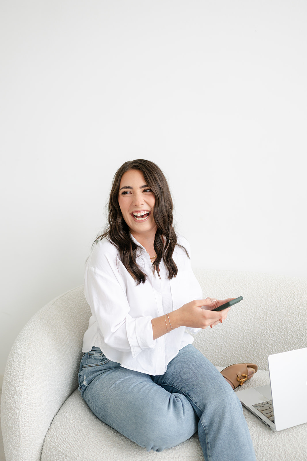 Woman sitting on a sofa, smiling, holding a smartphone, with a white background.