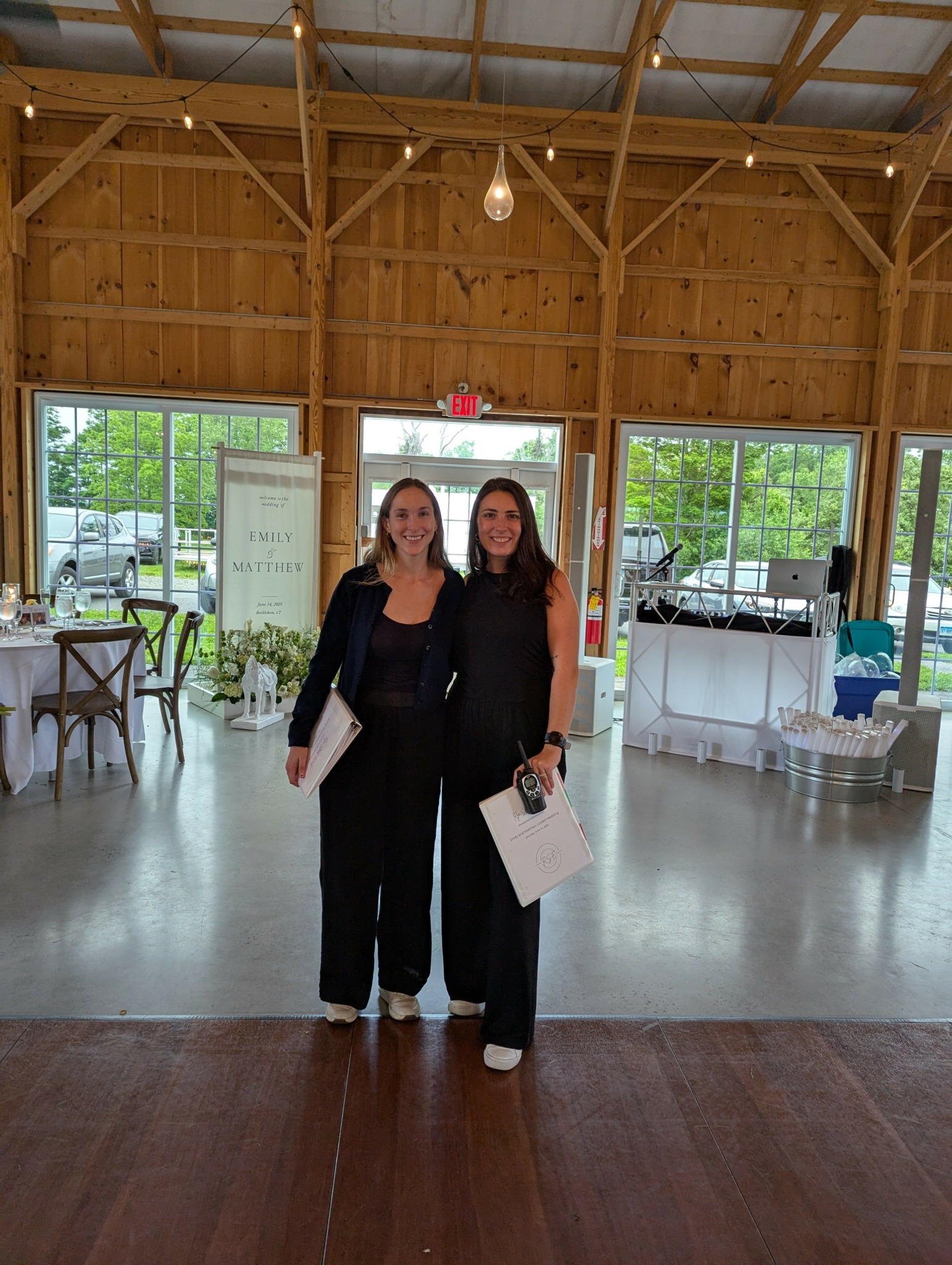 Two women standing inside a wooden barn with large windows, smiling and holding papers or bags.