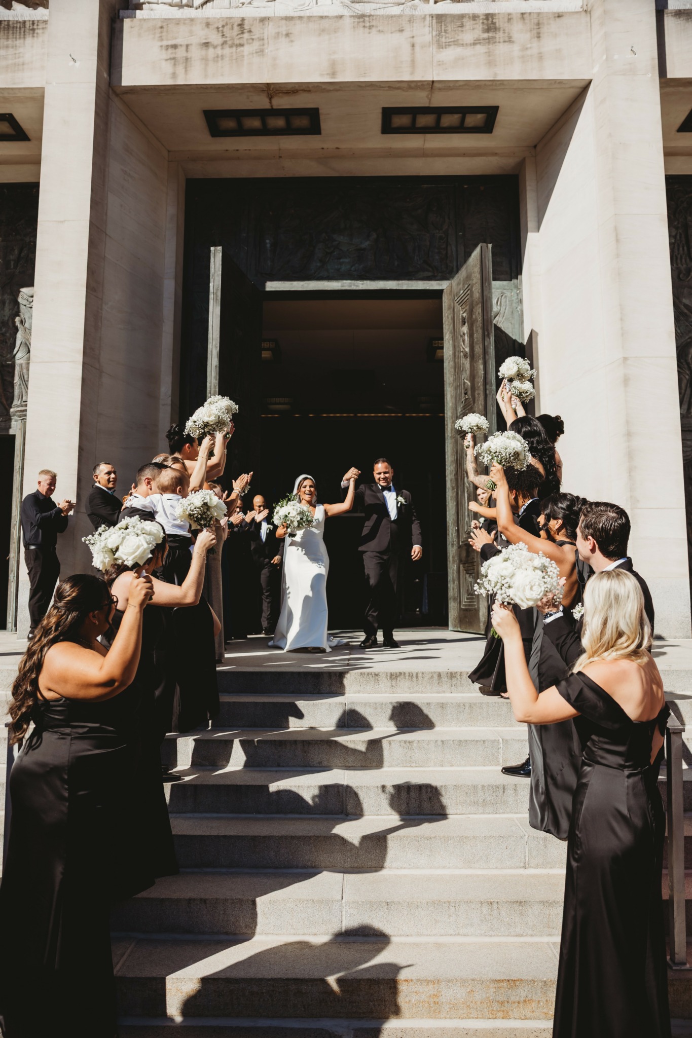 Group of people on steps outside building, some holding bouquets, celebrating wedding, with a bride and groom in center.