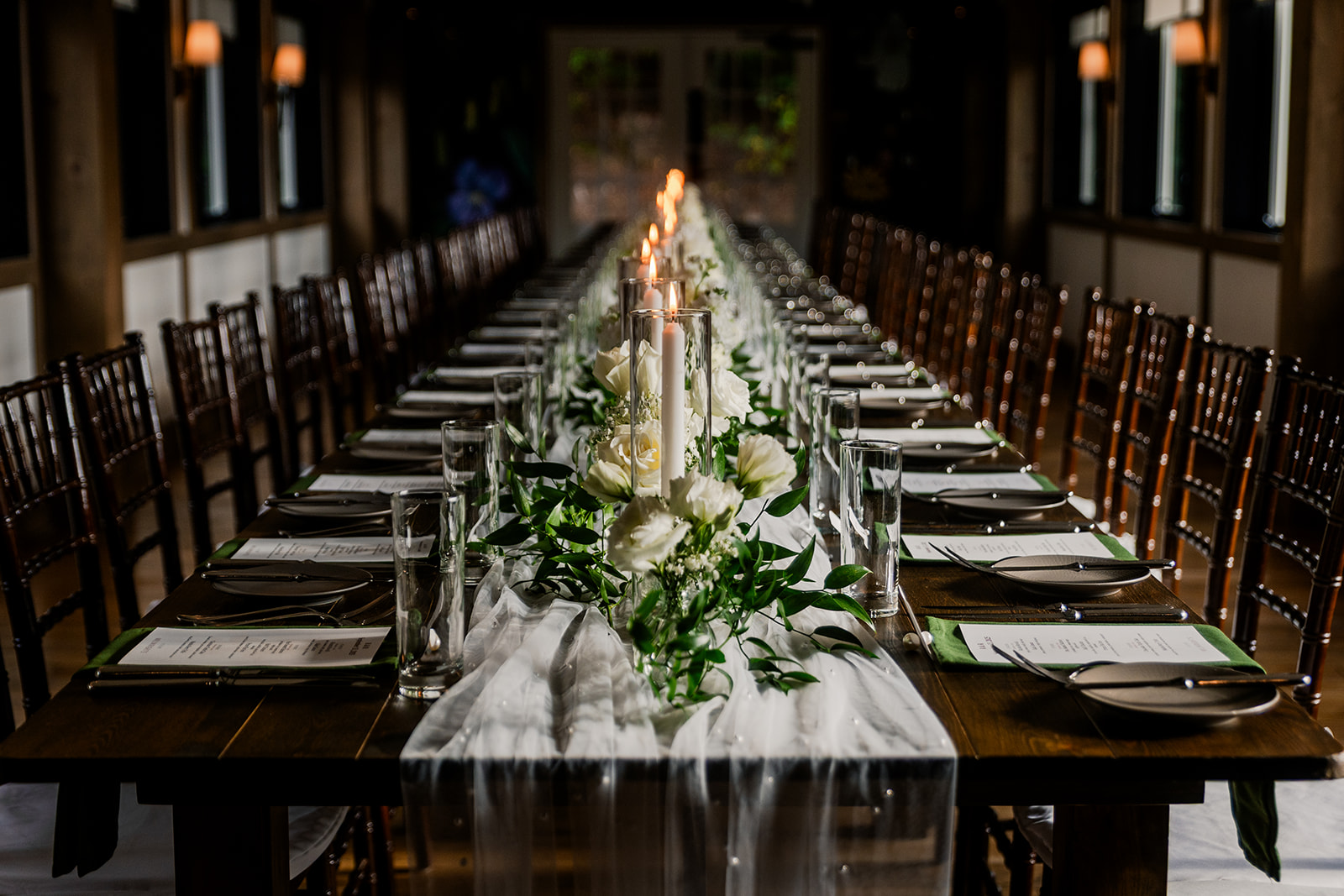 Long dining table set with white flowers, candles, glasses, and plates in a dimly lit room.