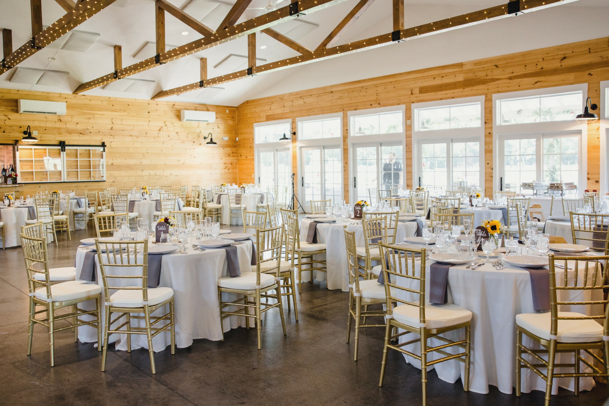 Event space with round tables covered in white tablecloths, gold chairs, large windows, and wooden walls.