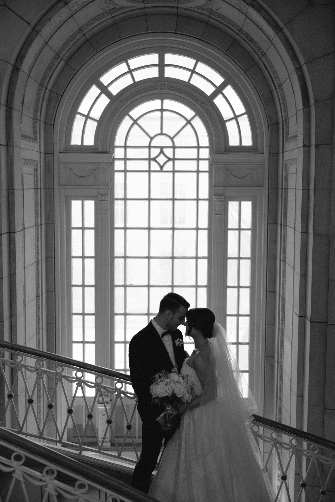 Silhouette of a bride and groom standing on a staircase in front of a large arched window.