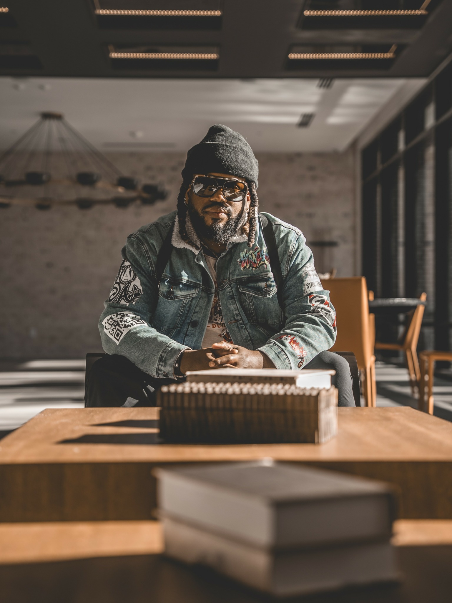Man with glasses, beanie, and beard sitting at a table in a modern room with large windows.