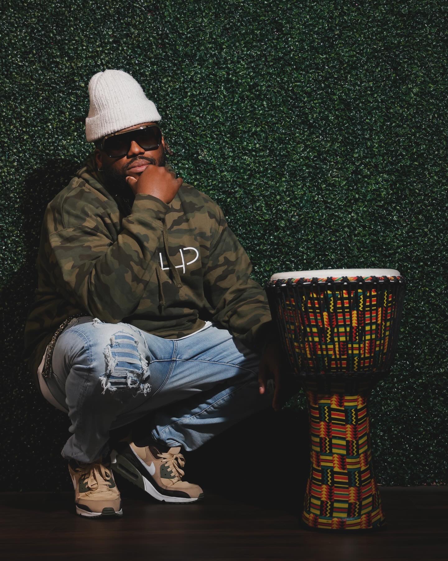 Person crouching next to a colorful drum, wearing a white beanie, sunglasses, camouflage jacket, and ripped jeans.