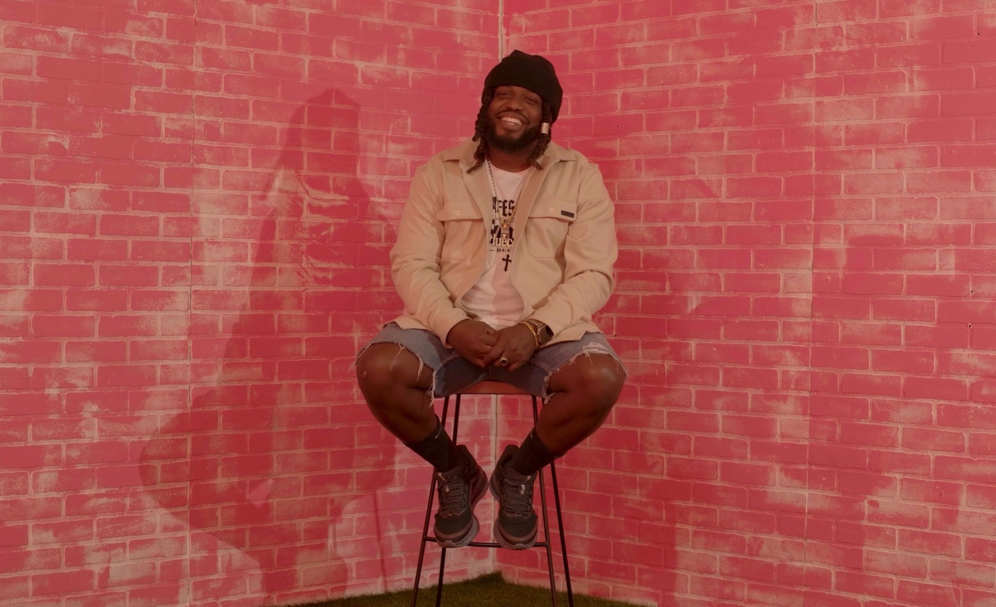 Person sitting on a stool against a pink brick wall, smiling, wearing a beige jacket, black beanie, and shorts.