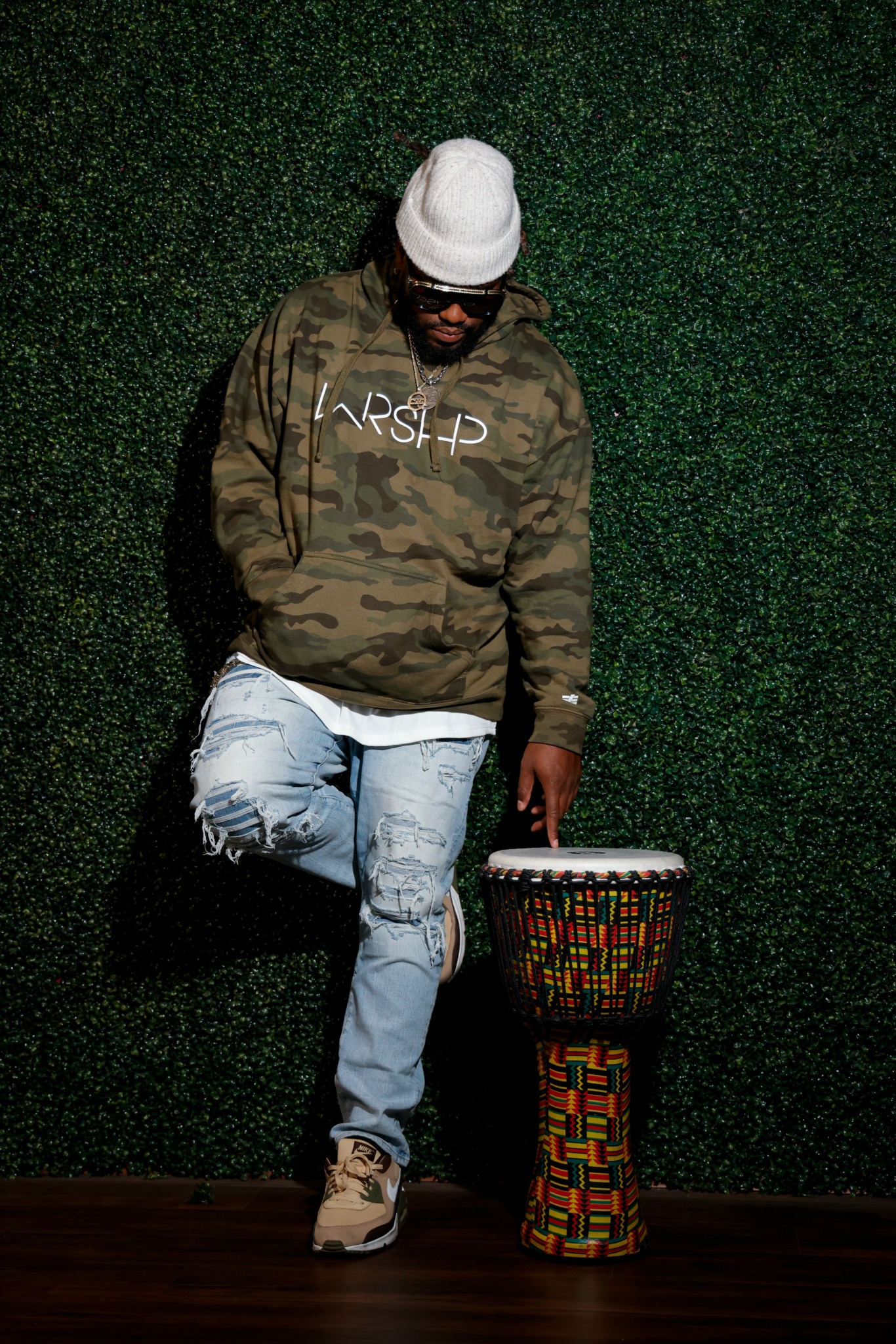 Person wearing a white beanie, camouflage jacket, and ripped jeans, standing next to a colorful drum against a green wall.