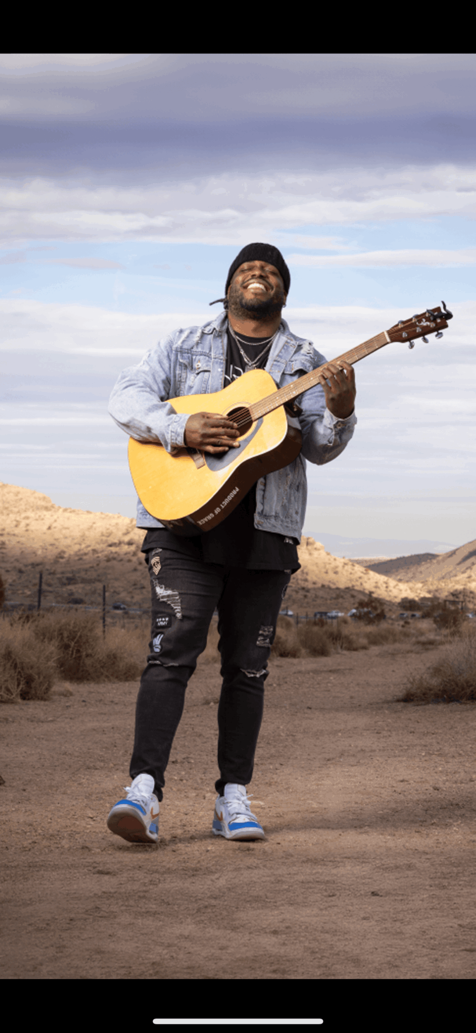 Person standing outdoors playing an acoustic guitar, wearing a denim jacket, black pants, and sneakers, with a cloudy sky background.