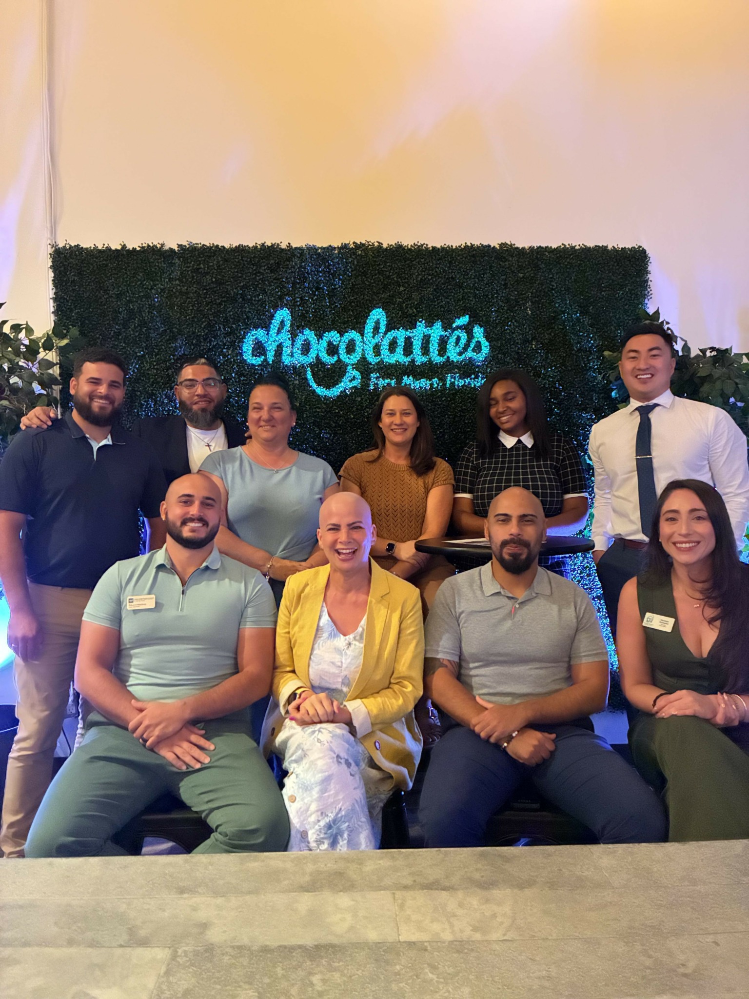 Group of ten people posing indoors in front of a green hedge with a sign that reads 'chocolatés'.