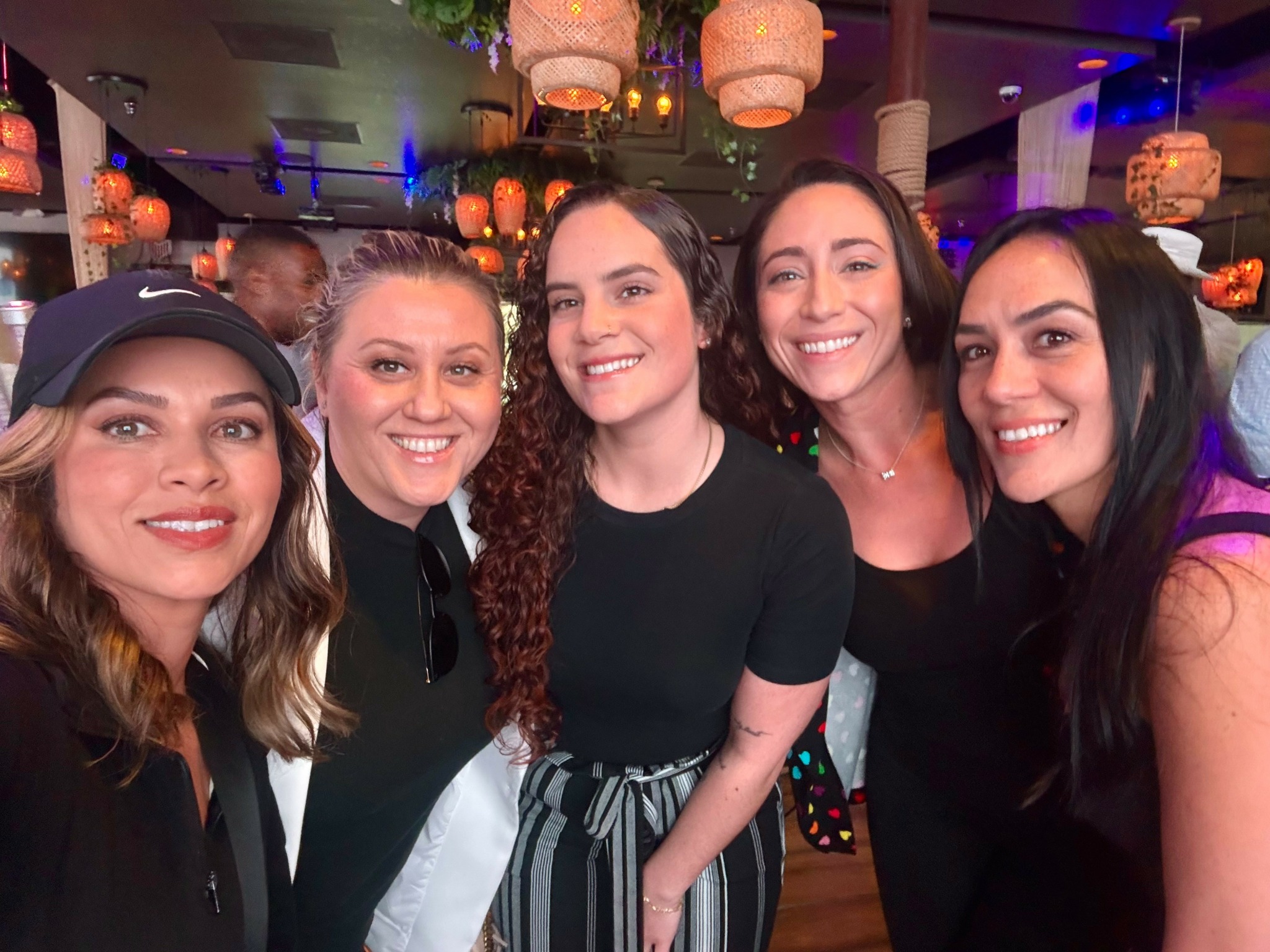 Five women smiling together in a lively indoor setting with hanging lights and colorful background.