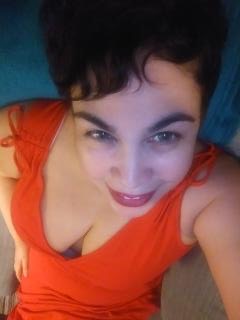 Woman lying down with dark hair, wearing an orange top, looking at the camera.
