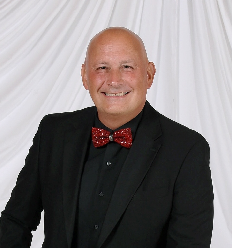 Bald man in black suit and red bow tie smiling, white draped background.