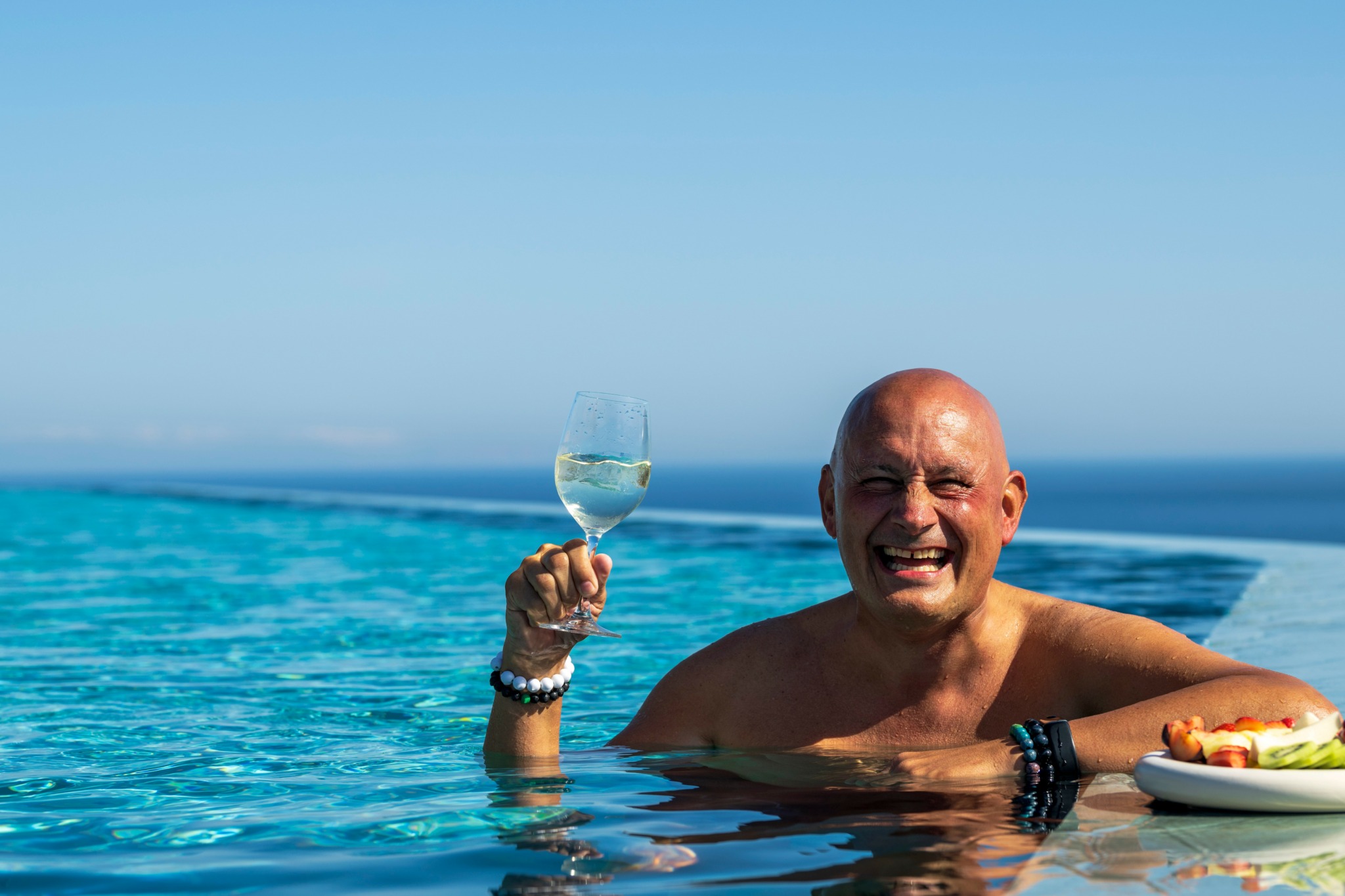 Bald man smiling in swimming pool holding a glass of water, with a plate of food nearby, against a blue sky.