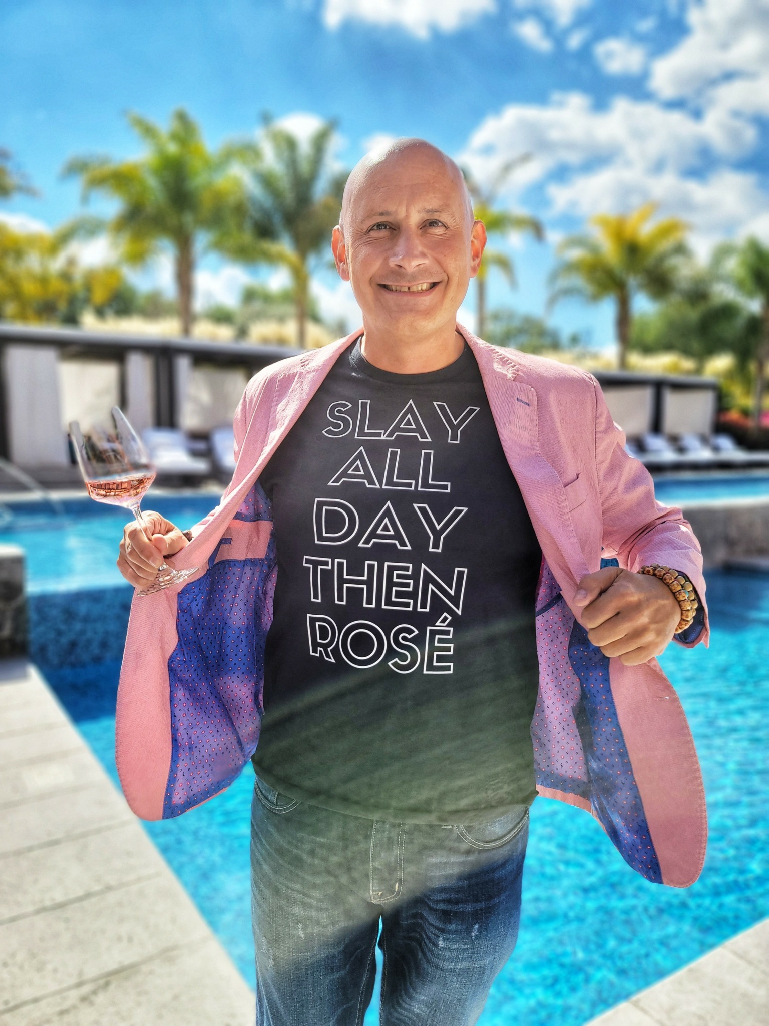 Man smiling, holding a glass of rosé wine, standing outdoors near a pool with palm trees and blue sky.