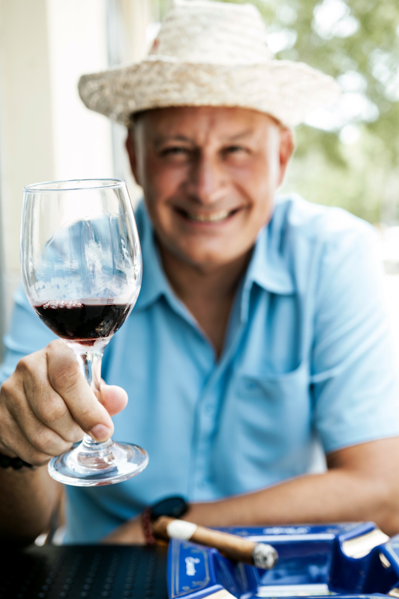 Smiling man in a hat holding a glass of red wine outdoors.