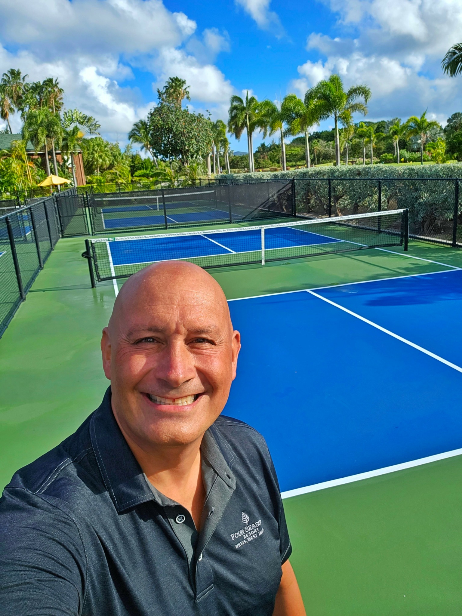 Man smiling in front of tennis courts with trees and blue sky in background.