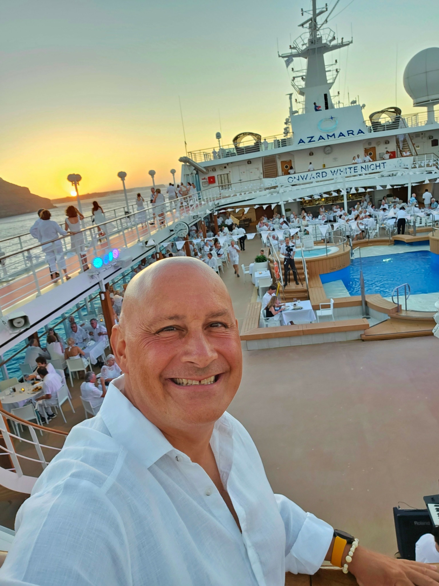 Smiling man in white shirt on cruise ship deck with pool and sunset in background.