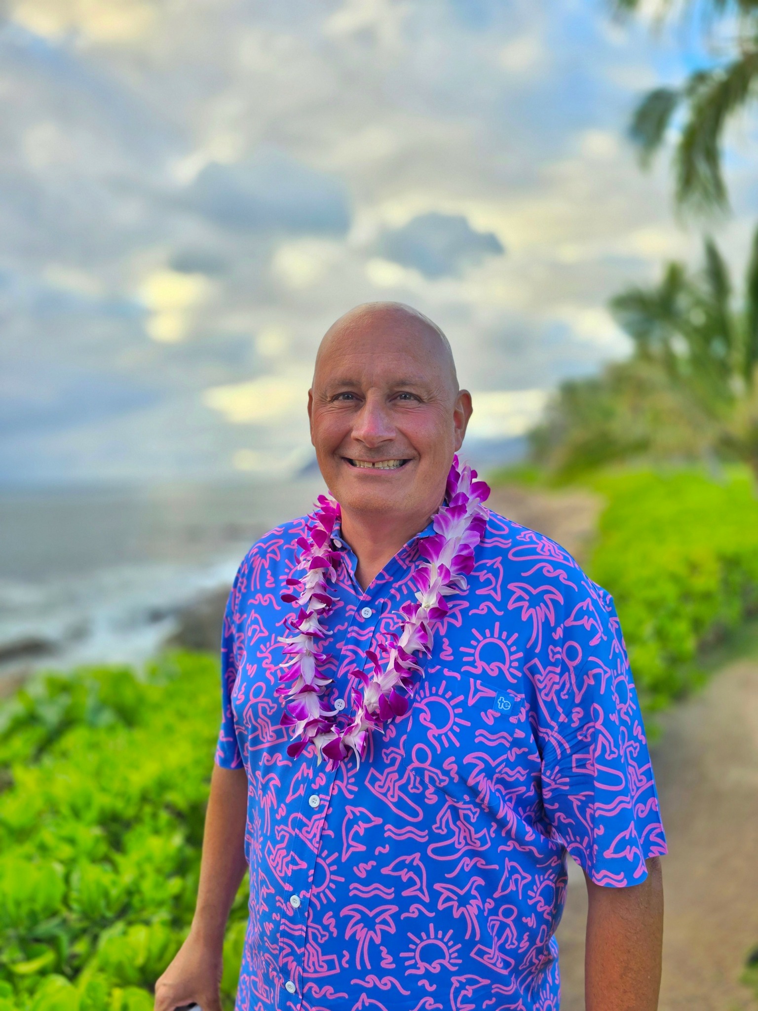 Smiling man wearing a blue Hawaiian shirt and a lei, standing on a beach with greenery and cloudy sky.
