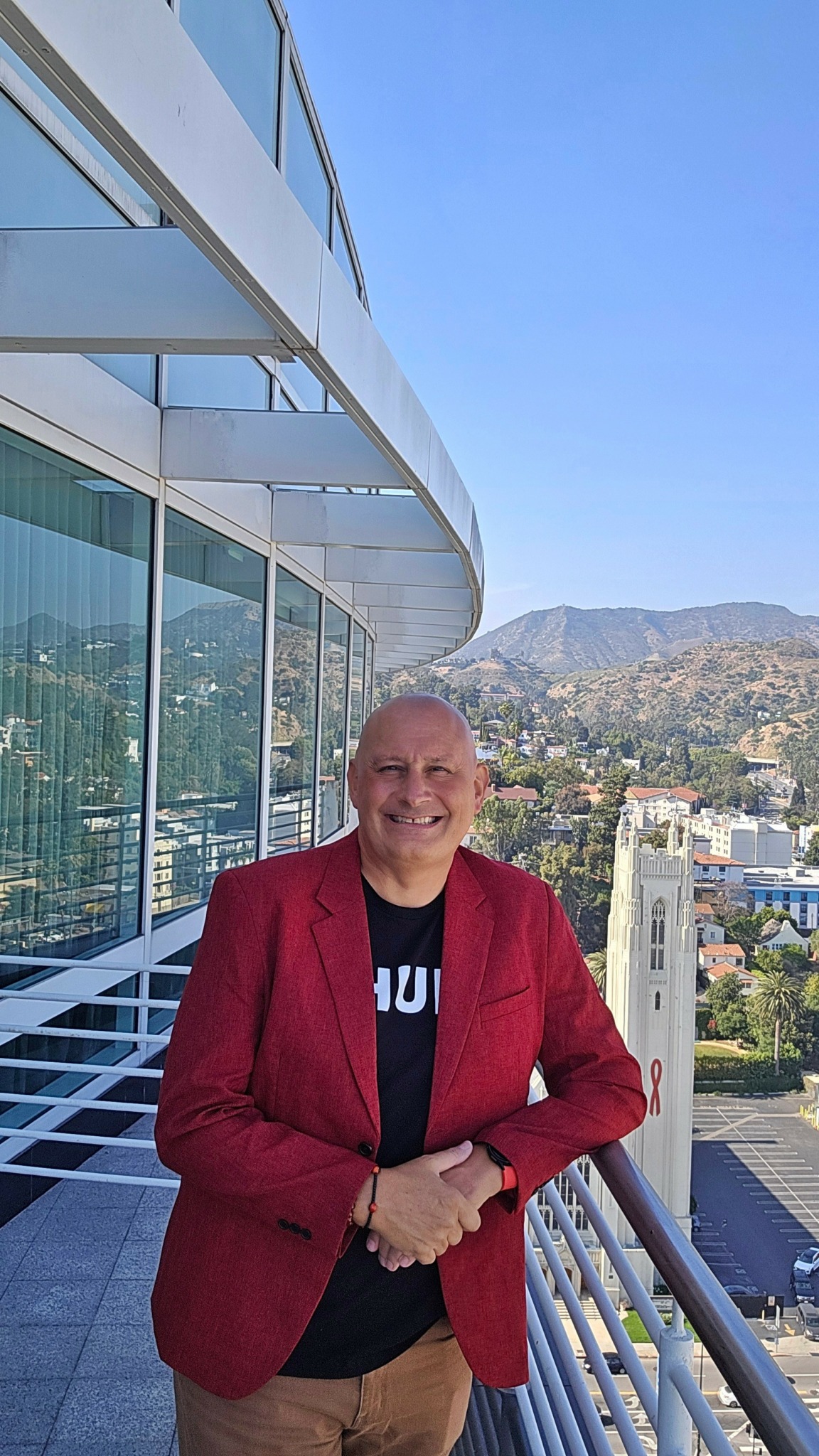 Smiling man in red blazer standing on balcony with cityscape and mountains in background.