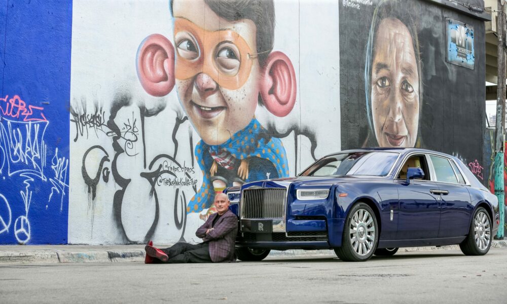 Person sitting on the ground next to a blue car in front of graffiti murals of cartoon and realistic faces.