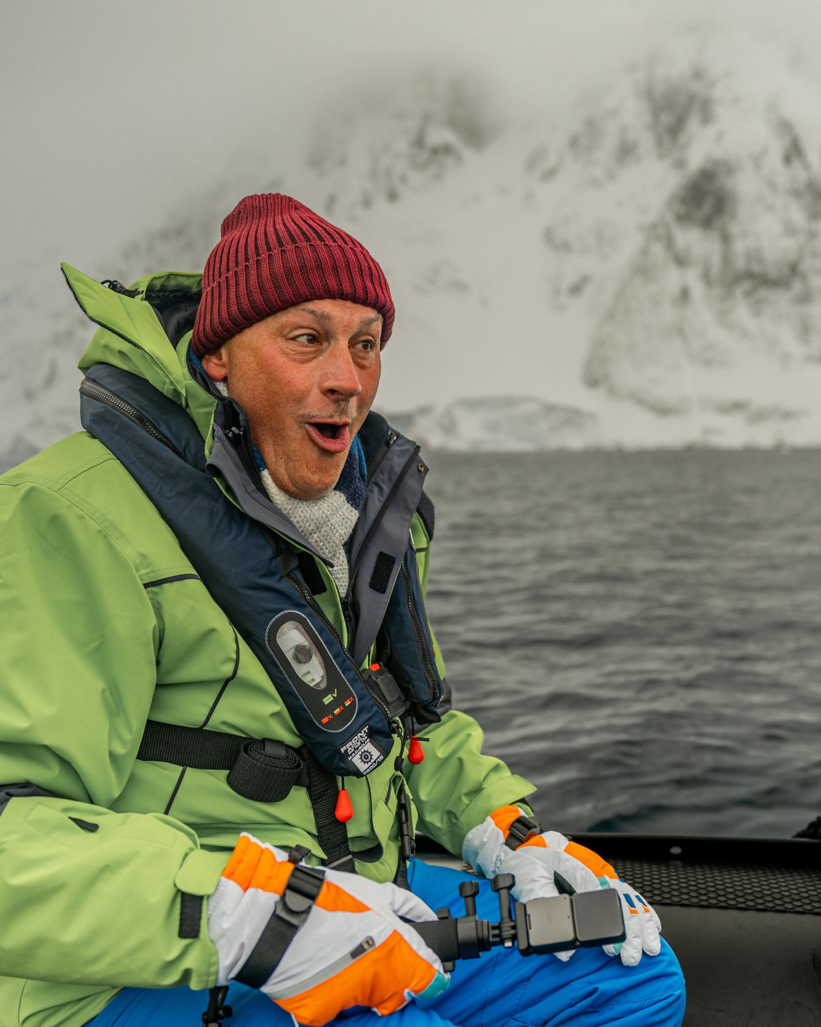 Man in green jacket and red beanie on boat, surprised, snowy mountains in background.