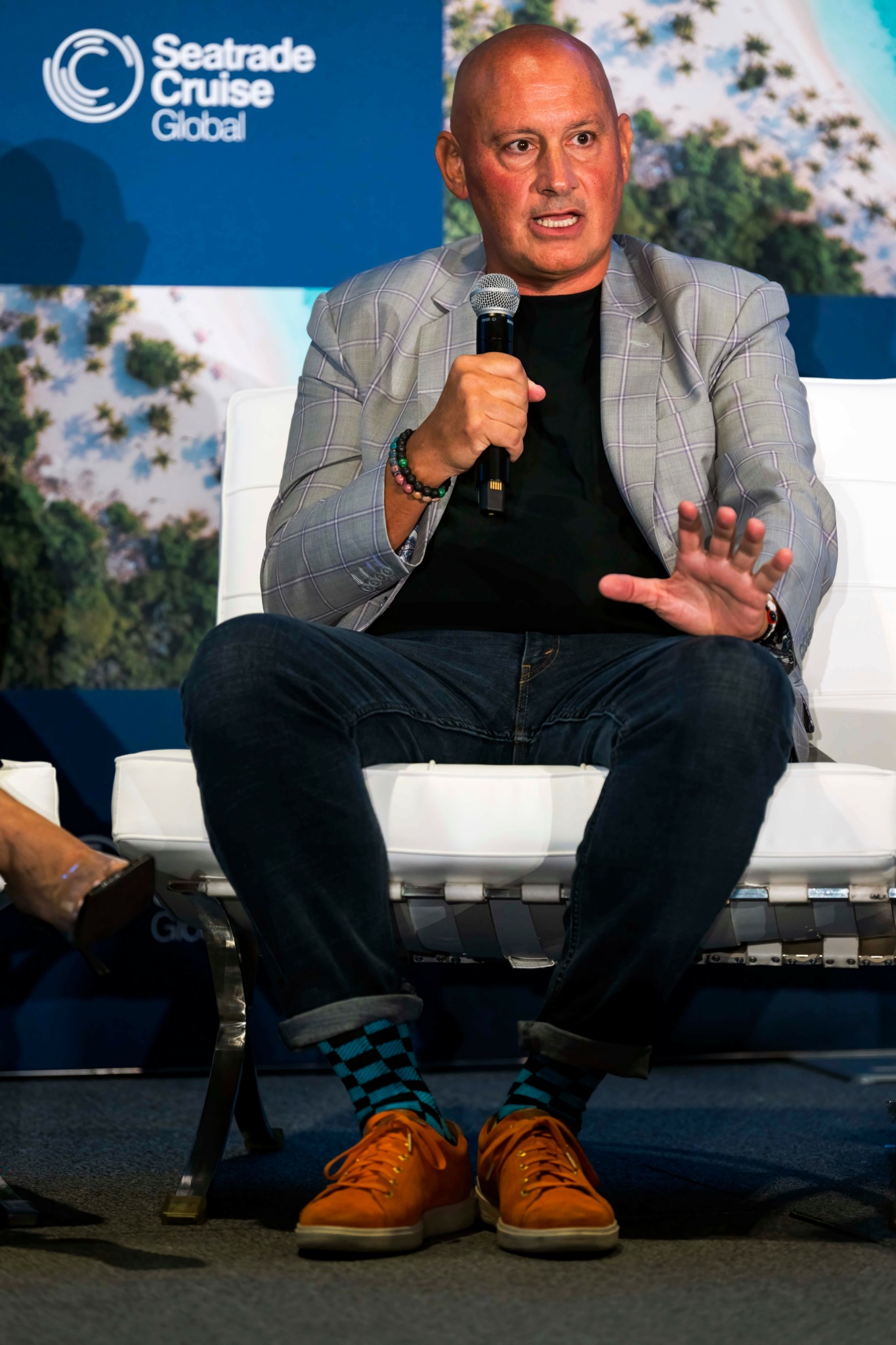 Man in gray blazer and black shirt speaking into a microphone, seated on a white chair, with Seatrade Cruise Global background.