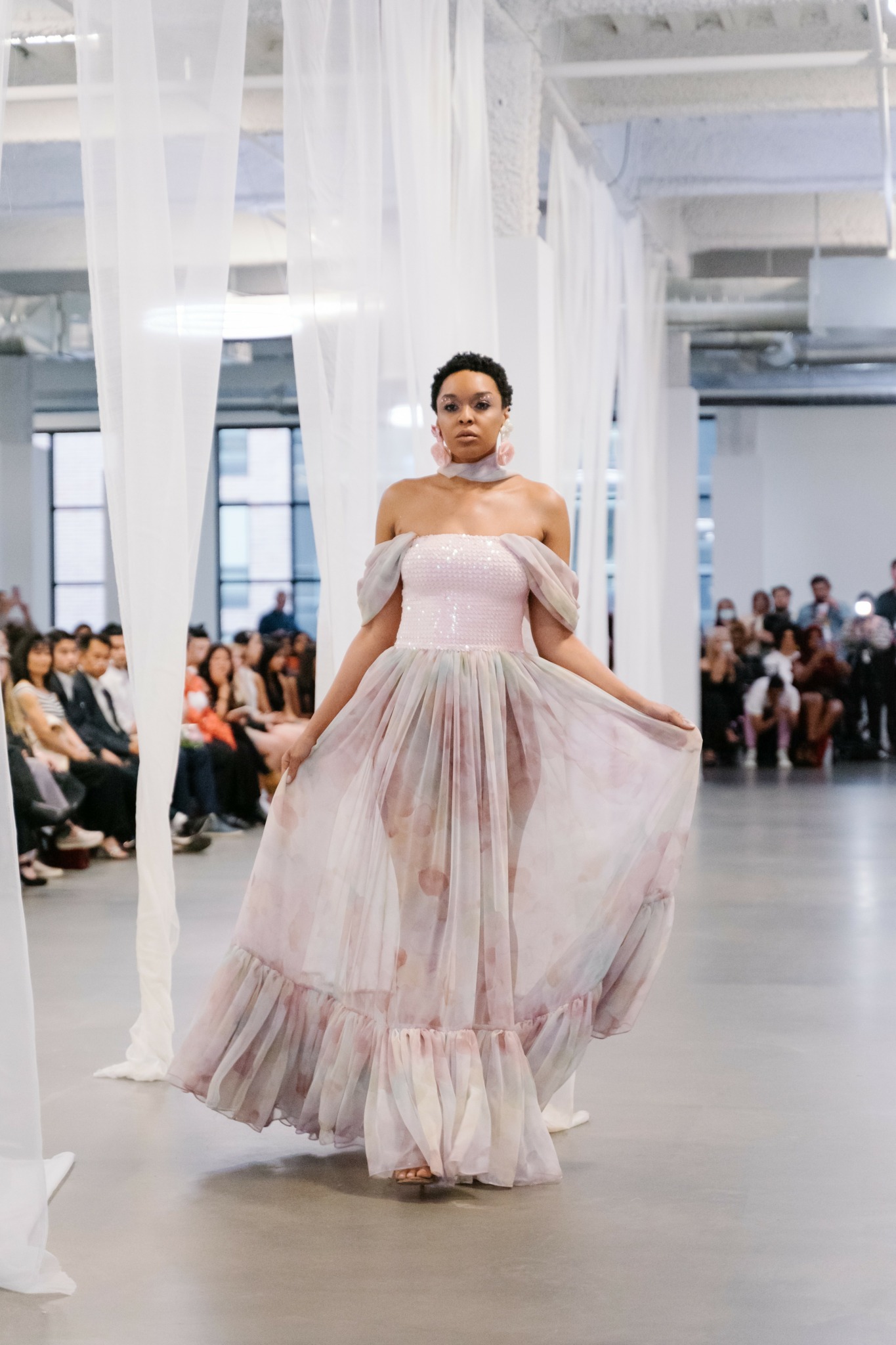 Woman in a pink strapless dress walking in a large, bright space with seated audience on both sides.