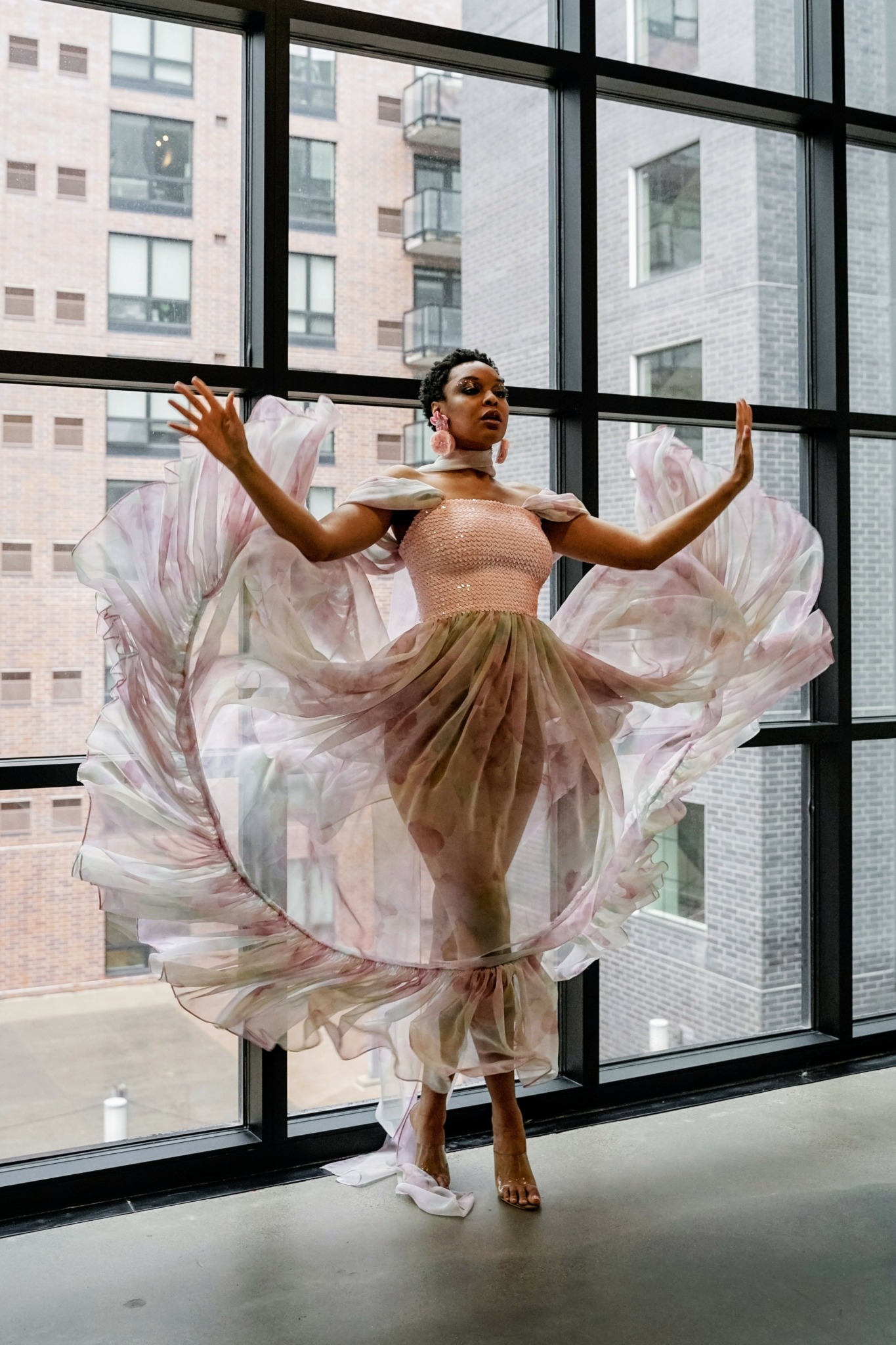 A dancer in a pink top and flowing skirt performs in front of large window panes with city buildings outside.